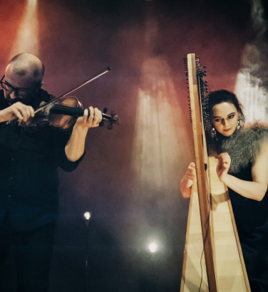 A woman and man are playing harp and violin respectively. They stage against an active background of smoke and coloured stage lighting streaming through.
