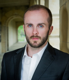 Tenor Thomas Walker stands in the hallway of an old building, leaning against a wall, looking directly into the camera.