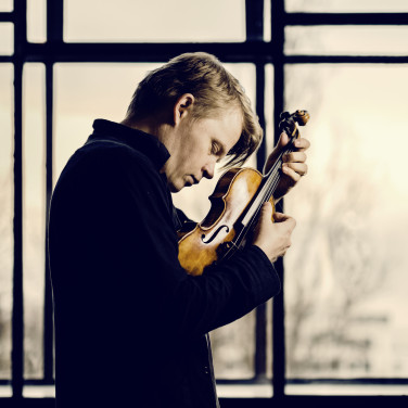 Violinist Pekka Kuusisto, eyes closed, holds his violin close to his face, plucking the strings with his fingers, as he looks down.
