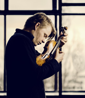 Violinist Pekka Kuusisto, eyes closed, holds his violin close to his face, plucking the strings with his fingers, as he looks down.