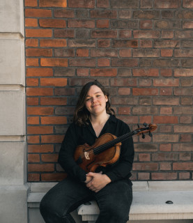 Francesca sits on a bench with a red brick wall behind her. She holds her viola and wears a black top and black trousers.