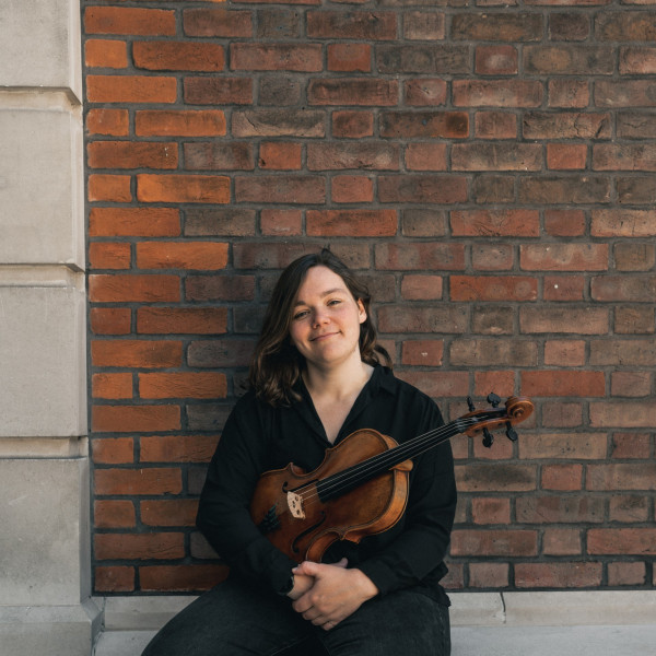 Francesca sits on a bench with a red brick wall behind her. She holds her viola and wears a black top and black trousers.