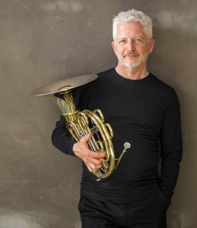 Ken holds his horn. He wear a black jumper.