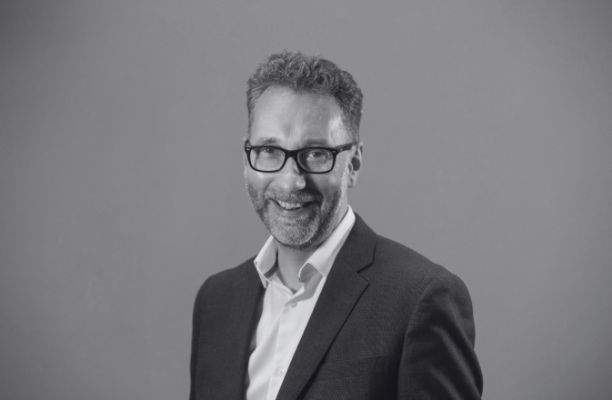 A black and white image of Gavin Reid. He wears glasses and a black blazer.