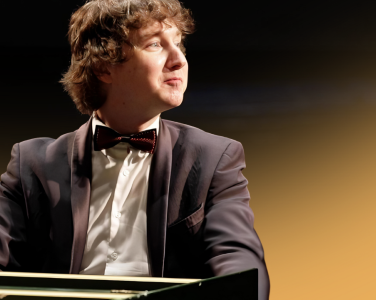 Maxim Emelyanychev sits at the keys. He smiles off to his left. He wears a black suit and bowtie.