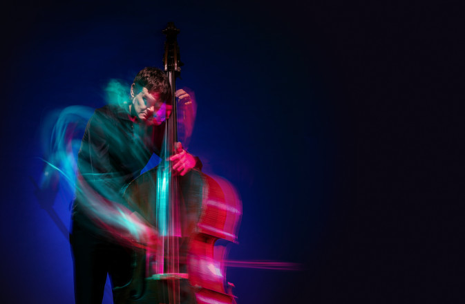 SCO Double bassist Nikita Naumov stands in front of a dark background playing his bass. His movements are highlighted with neon pink and turquoise swirls.