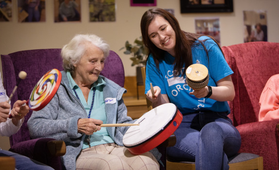 An SCO Creative Learning team member in a blue SCO tshirt holds a small bongo as an older lady smiles playing a handheld drum in the chair next to her.