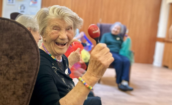 An older lady in an armchair smiles with a maraca in her hand.