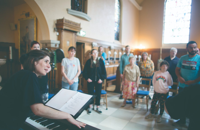 Moira leads a Voices session in a church. She is sat smiling behind a piano as people circle around her.