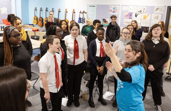 SCO Voices Schools Choir sing with red ties as leader Moira in a blue SCO t-shirt stands in front with her arm in the air.