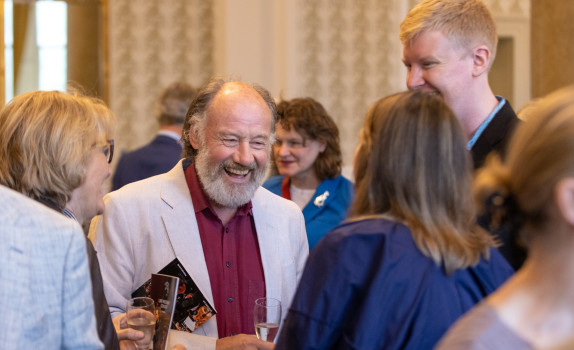 An SCO donor smiles during a reception. He has a grey beard and wears an off white blazer with a maroon shirt.