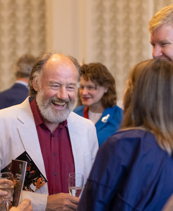 An SCO donor smiles during a reception. He has a grey beard and wears an off white blazer with a maroon shirt.