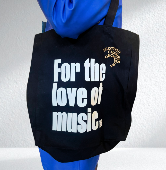 A person holds a black tote bag featuring the SCO logo in orange and For the Love of Music displayed in white text.