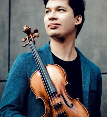 A man stands against a black background made up of four tiles. He looks off to his right and is holding a violin at its base. Its front faces the camera.
