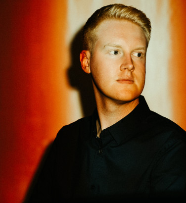 Against a red background, a man in a dark jacket and shirt looks out of frame. His face is illuminated by a light from above.