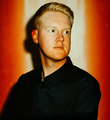 Against a red background, a man in a dark jacket and shirt looks out of frame. His face is illuminated by a light from above.