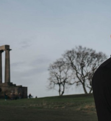 A man stands in profile (facing the camera) on a hillside, framed by two trees, with a series of stone columns from an unfinished temple on the far left