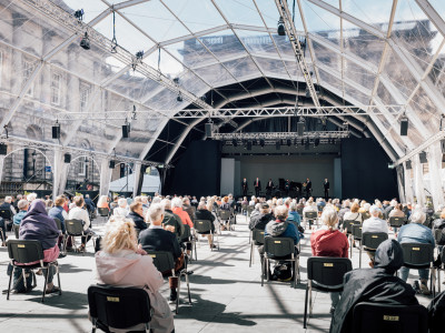 An audience sit in a glass marquee facing away from the camera towards a stage where musicians sit with instruments.