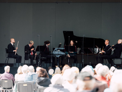 An audience face away from the camera looking towards a stage where an ensemble of musicians sit with different instruments.