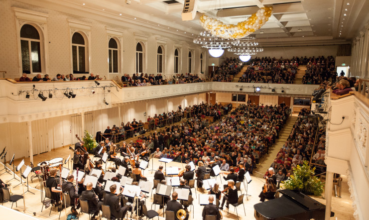 The orchestra take their seats on stage in front of the audience of a sold out concert.