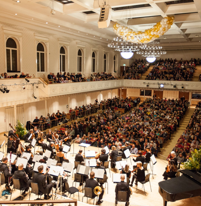 The orchestra take their seats on stage in front of the audience of a sold out concert.
